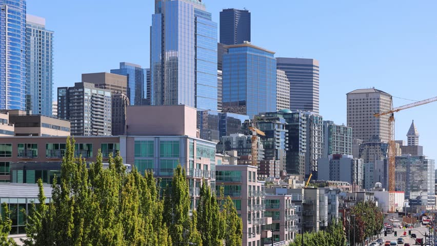 Aerial view of the Downtown Seattle skyline featuring modern glass skyscrapers, construction cranes, and lush green trees under a clear blue sky in Seattle, Washington, United States.