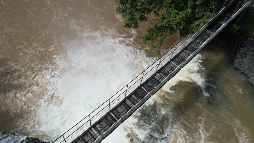 Aerial drone footage capturing dramatic waterfall cascades beneath rustic wooden suspension bridge flowing river water creates energetic motion landscape scene outdoor nature scenery.