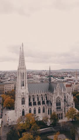 Spinning aerial view of a cathedral featuring twin spires, surrounded by a vibrant city landscape, showcasing architectural beauty in Vienna at the Votivkirche church in vertical video format.