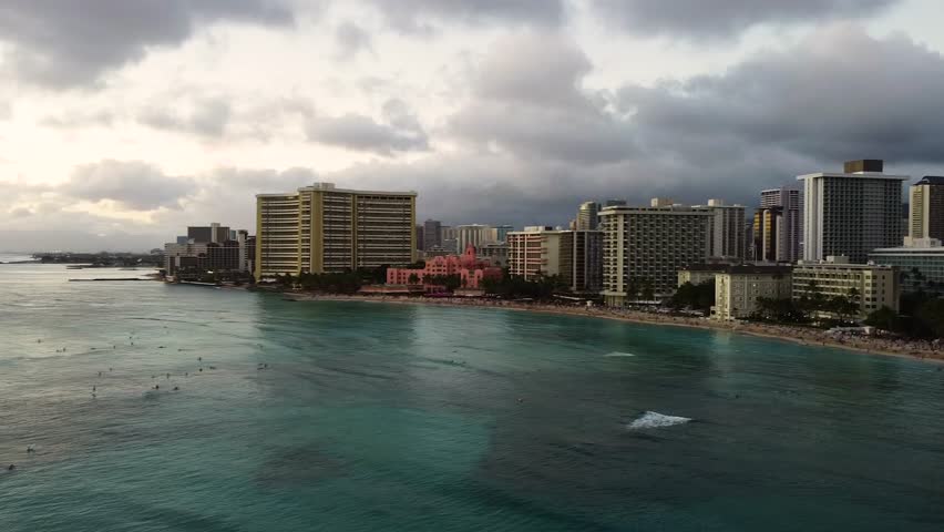 Panning aerial view of waikiki beach in honolulu, hawaii, showing hotels, skyscrapers, and surfers in the ocean at sunset