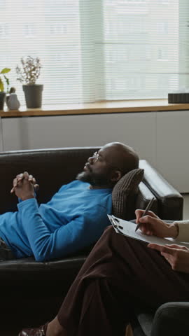 Vertical high angle shot of middle aged African American man lying on couch and talking during psychotherapy session while therapist taking notes. Perfect for mental health and wellness campaigns