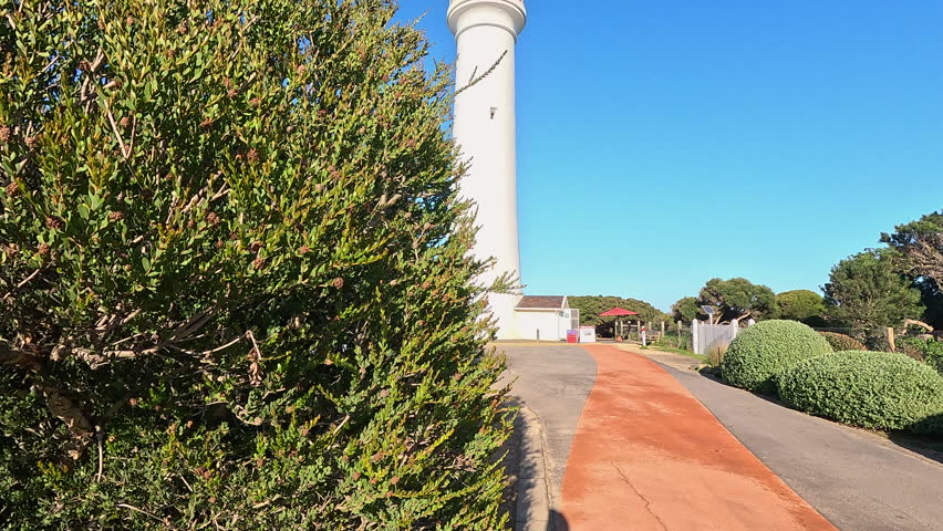 Tall white lighthouse with red top rises beside a curved path framed by dense green shrubs under clear blue sky.