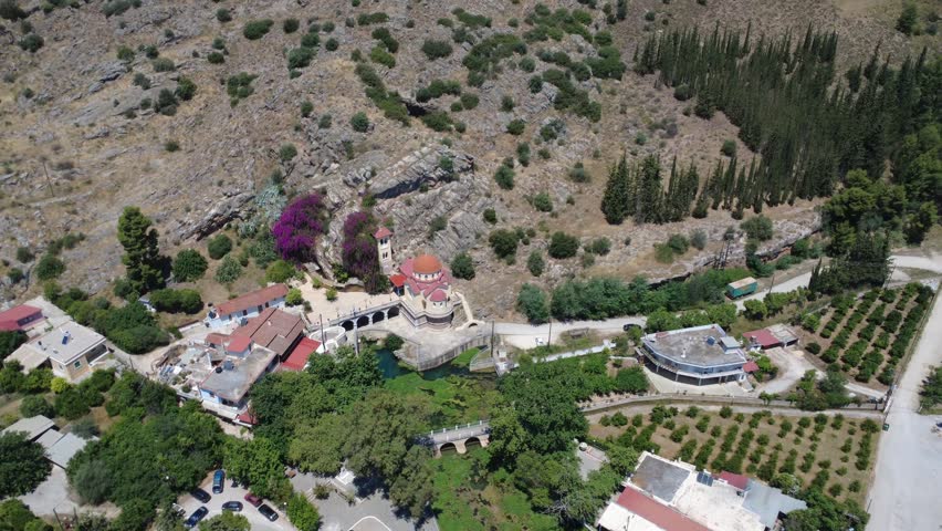 Panoramic aerial view reveals an Orthodox church in a green valley beside a rocky mountain, surrounded by trees, a lake and a bridge. Travel and recreation destination in Greece, Europe.