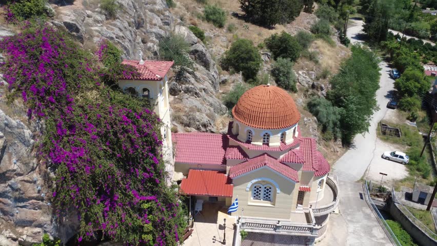 Aerial close wide angle view of a traditional Orthodox Christian church with a red dome, tall stone bell tower and blooming purple bougainvillea growing on rocky terrain in Greece, Europe.