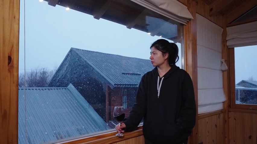 Woman holding red wine beside large window watching snowfall over mountain cabins in winter, swirling contemplating gaze