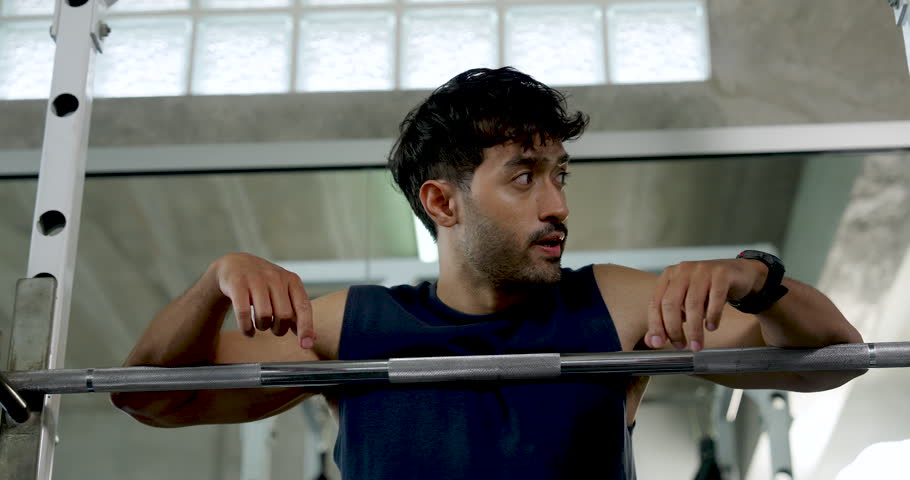 Young Asian man pauses at barbell rack during shoulder workout in modern gym, gripping weight bar and looking down between sets, solo strength training, recovery, wellness, healthy lifestyle.