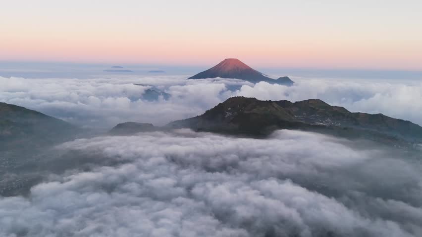 Aerial view of the volcanic peak of Mount Sindoro rising above a thick layer of clouds during sunrise in Indonesia.