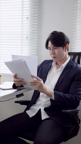 Professional Asian male freelancer in suit examining business documents while working at bright contemporary workspace