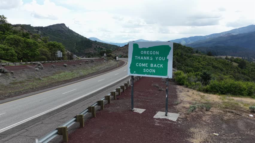 Oregon Thanks You Come Back Soon farewell sign stands roadside along a winding mountain highway flanked by rugged rocky outcrops and dry shrub hillsides in Oregon, USA. Aerial drone view.