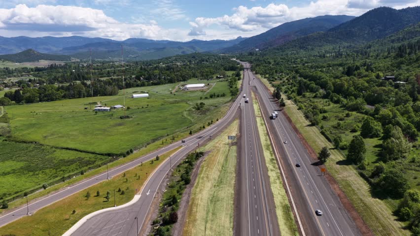Interstate highway with moving traffic slices through a broad lush green valley flanked by forested mountain ranges under dramatic partly cloudy skies in Oregon, USA. Aerial drone flyover.