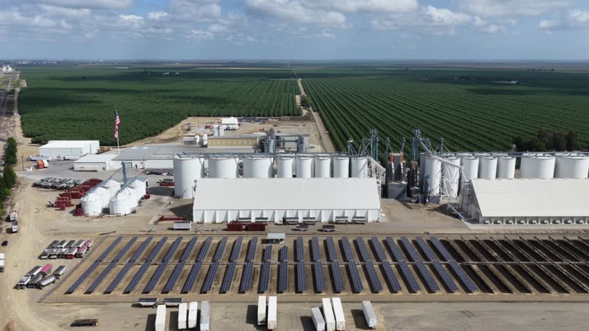 Large almond processing facility with grain silos, solar panels and warehouses surrounded by vast orchard rows stretching to the horizon in Arbuckle, California, USA. Aerial drone wide shot
