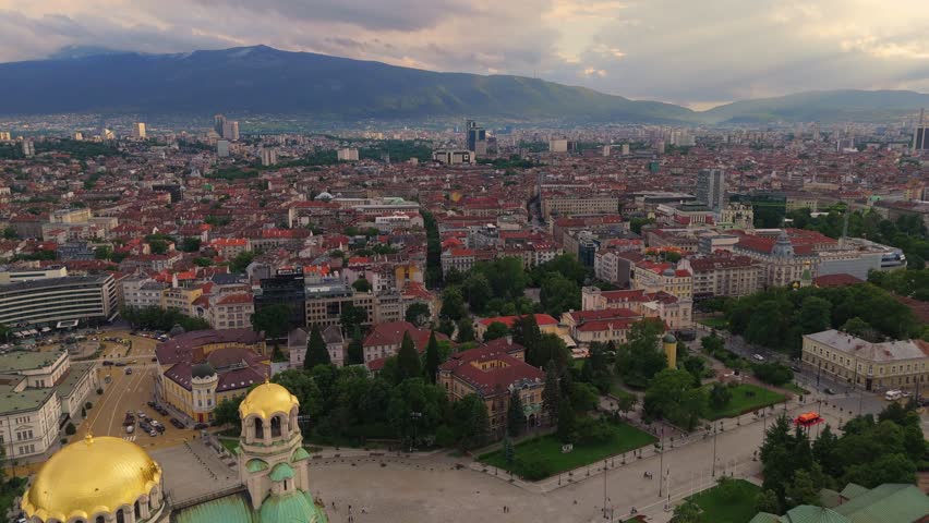 Drone perspective gliding past the shimmering golden domes of Alexander Nevsky Cathedral at sunset, showcasing the sprawling Sofia skyline Bulgaria capital city