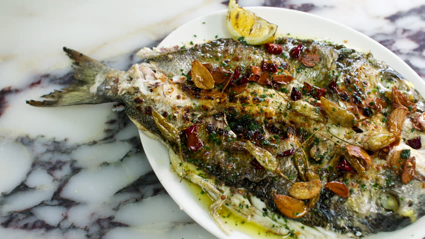 Whole grilled fish garnished with herbs, garlic, and lemon on a marble table surface