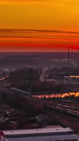 Aerial vertical timelapse of Jelgava railway station in Latvia with freight trains cargo wagons and river reflection under dramatic red sunset sky over misty town