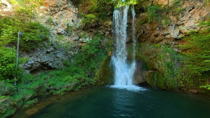 Water cascades down a rocky cliff into a deep pool. Lush greenery surrounds the waterfall