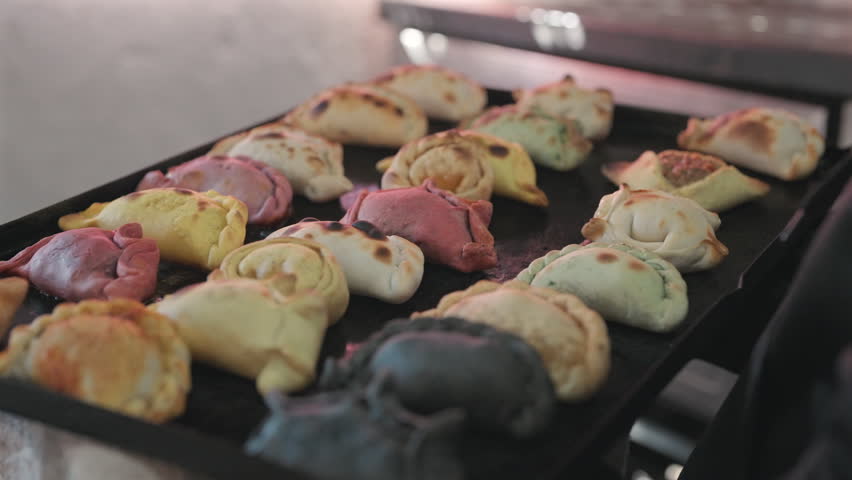 Freshly baked Argentine empanadas with assorted fillings rest on a baking tray, showcasing traditional cuisine, handmade preparation and colorful folded pastry shapes, close-up hanheld shot.