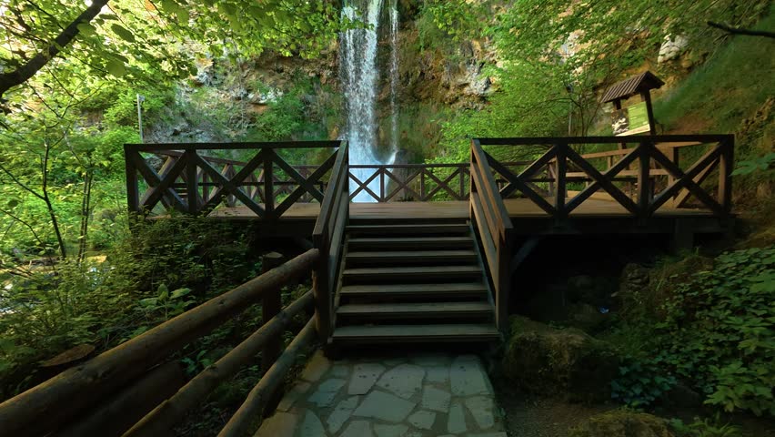 Lush green forest surrounds a tranquil waterfall. Wooden bridge invites viewers to approach the scene. Location: Waterfall Veliki Buk. Balkans, Serbia.