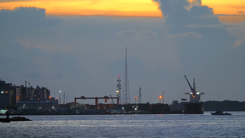 Twilight port view shows Male harbor cranes and city lights beneath a dramatic cloud bank with dark shoreline calm water and fading orange light over the Maldives at tropical dusk