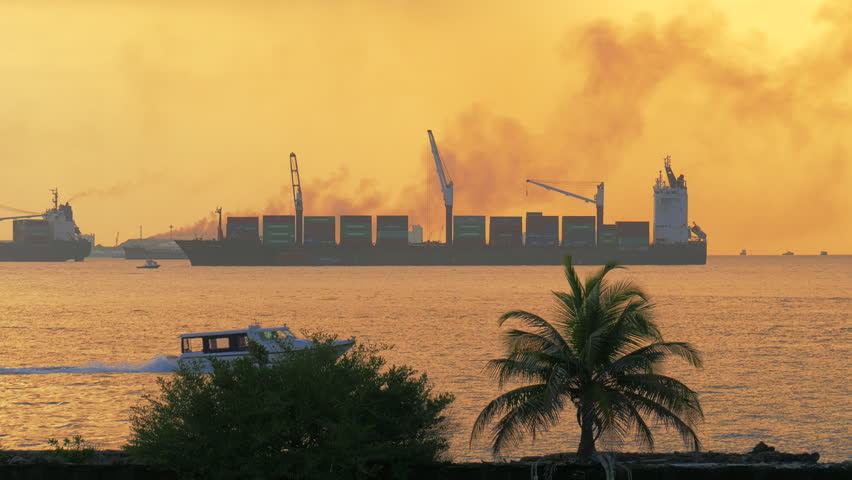 Golden hour harbor view a container ship at Male port with cranes smoke plume palm silhouette and small boats crossing the glowing water near the Maldives coast in evening light
