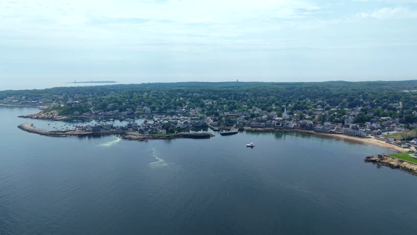 Rockport Massachusetts Bay with harbor village, granite coastline, sandy beach and boat wake on calm water, aerial push forward and tilt down