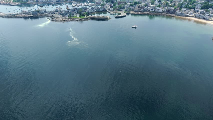 Rockport Massachusetts harbor village with anchored boats, granite coastline, church steeple, residential rooftops and a motorboat on calm water, aerial push forward and tilt up
