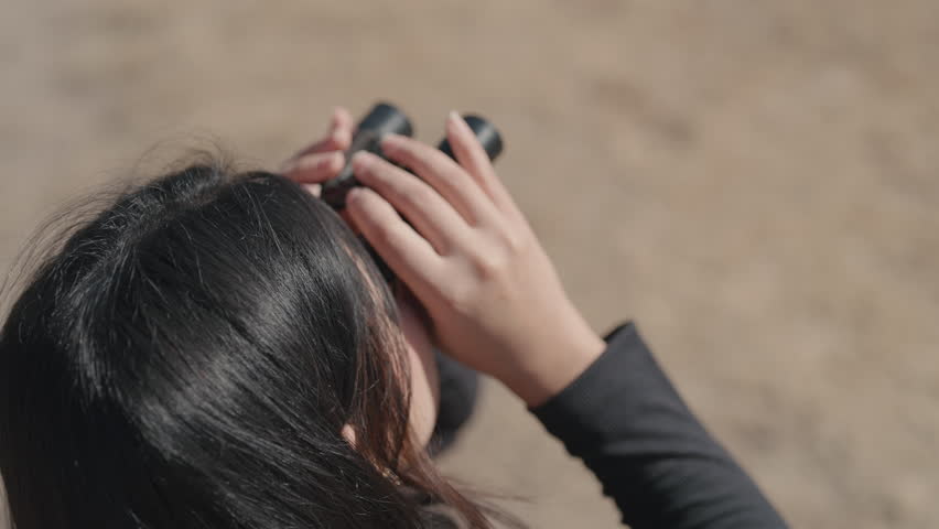 Close-up of a woman using binoculars while observing a remote mountain landscape.