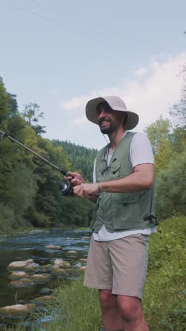 vertical Joyful fisherman wearing a hat and vest reels in his fishing rod and celebrates his catch with a happy fist pump while standing on the green bank of a beautiful river surrounded by nature