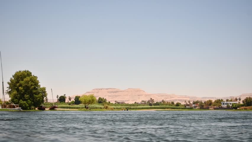 Boat ride along Nile River shows green shore, village houses and desert hills beneath wide Egyptian sky on warm sunny day.
