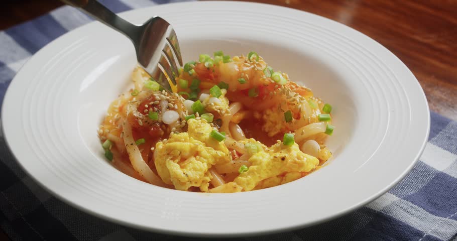 Close up of homemade shrimp udon noodles with scrambled eggs green onions and sesame seeds served in white bowl.A fork picks up the freshly cooked noodle dish prepared with shrimp vegetables and eggs