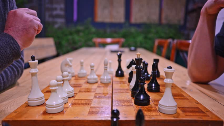 Two people play chess on wooden board outdoors. Hands move pieces during game