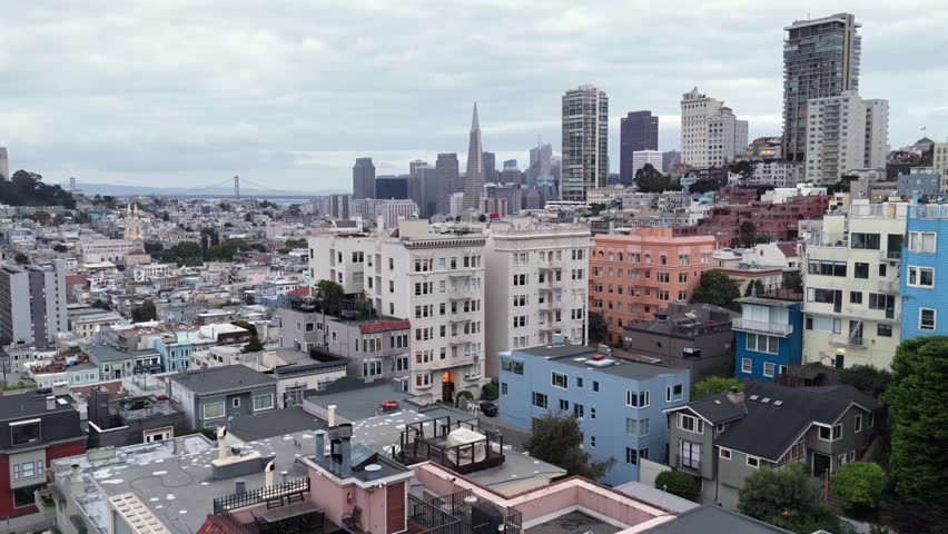 Colorful pastel apartment facades lining steep Lombard Street hill with Transamerica Pyramid and dense San Francisco skyline beyond in San Francisco, California, USA. Aerial drone backwards shot.