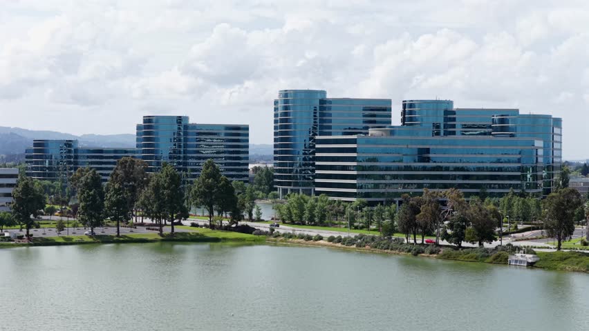 Oracle headquarters campus with iconic blue glass towers reflected in lagoon, tree-lined waterfront promenade and rolling hills in distance in Redwood City, California, USA. Aerial drone wide shot