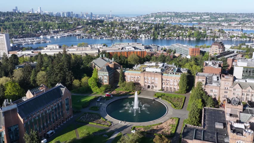 Historic University of Washington campus fountain, brick buildings and lush grounds overlooking Lake Union with Space Needle and skyline in distance. Seattle, Washington, USA. Aerial drone wide shot.
