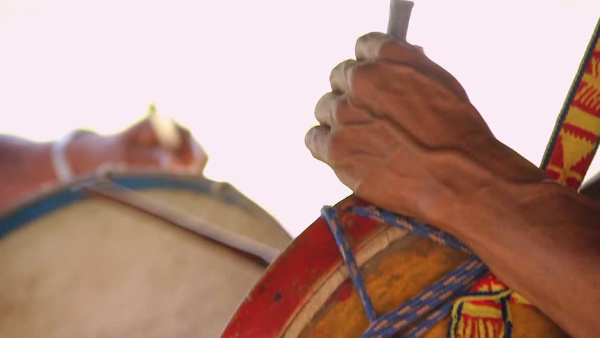 Hands beat drum with rhythm and passion. Drum skin taut, painted with bold colors. Cultural celebration pulses through steady beats. Sunlight glows softly behind musicians. Lamas, Tarapoto. Peru.