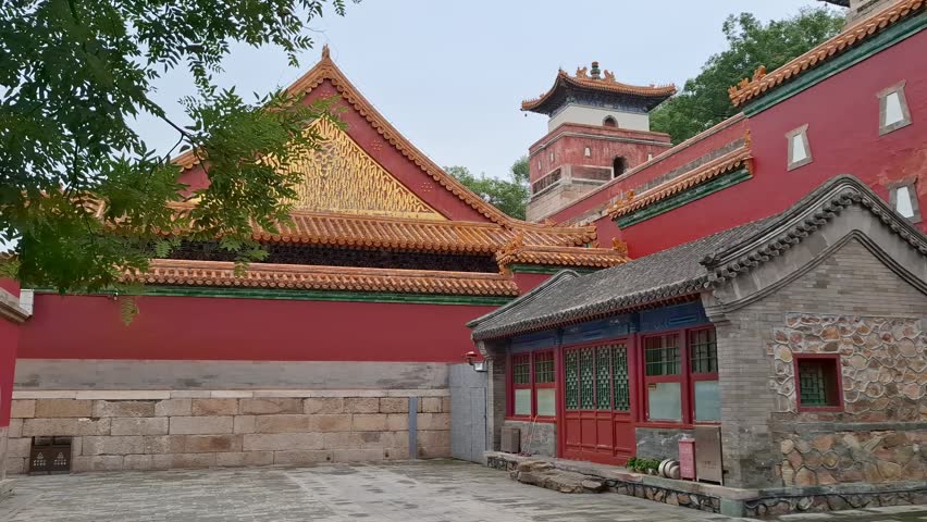 Beautiful courtyard inside Summer Palace temple complex in Beijing featuring detailed ornate rooftops and building. Green tree branches decorate foreground of ancient landmark. Rich asian history