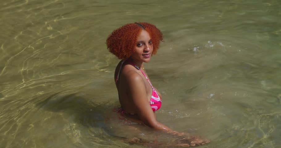 A girl in a stylish bikini its in the surf of a tropical paradise, the translucent blue water swirling around her ankles.