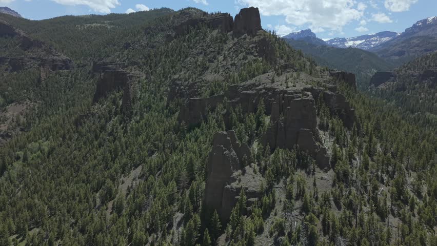 Aerial view of rhyolite cliffs and forested canyon terrain in Shoshone Canyon, Wyoming, with rugged rock formations, pine-covered slopes, and snow-capped mountains in the distance.
