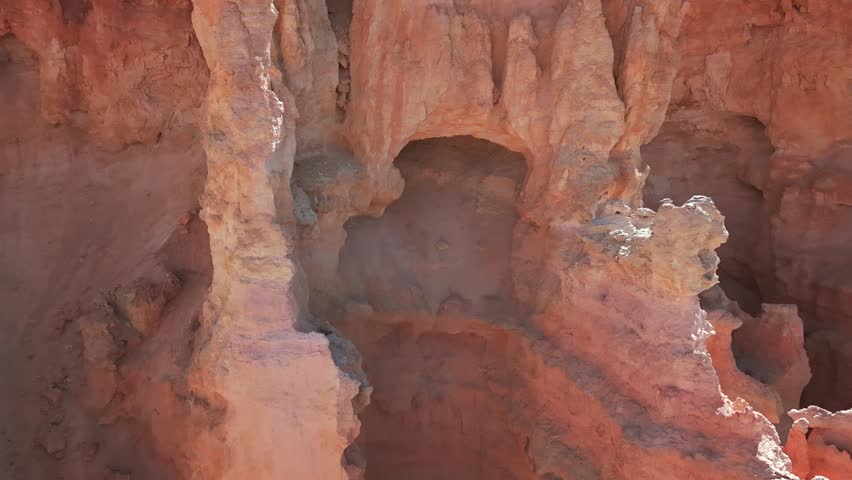 Aerial drone footage pulling away from a cave-like opening in a limestone cliff near Bryce Canyon, Utah. The shot reveals rugged rock formations, pine trees along the cliff top, and a bird flying through the canyon landscape, captured in 4K resolution.