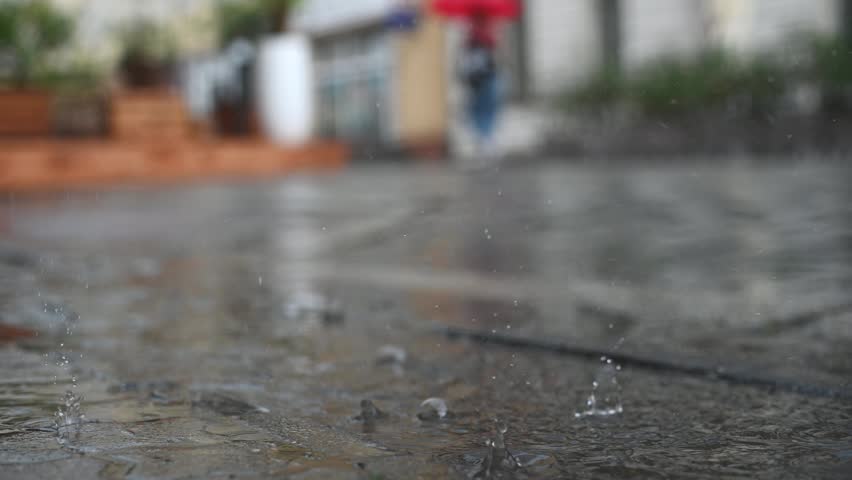 Raindrops Splatter On City Cobblestones As A Person Walks Under An Umbrella In The Background