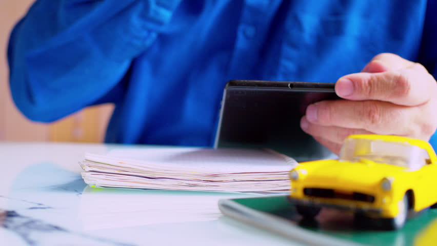 Businessman using tablet and writing notes in notepad at desk. Focus on hands, yellow toy car on table. Close up, shallow depth of field, natural daylight. Modern autobusiness data tracking.
