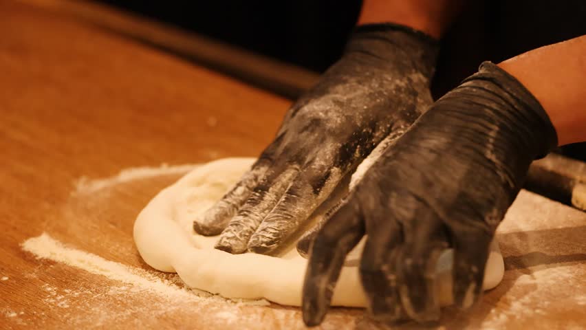 Artisan baker kneads and stretches fresh pizza dough on floured wooden surface under warm lighting.