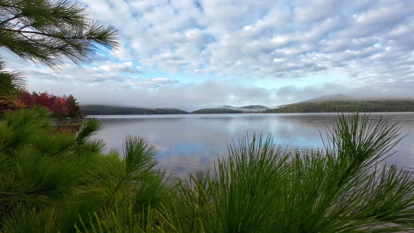 Cinematic view of calm water reflecting clouds and colorful autumn trees on Grand Lake in Achray, Algonquin Provincial Park, Ontario, Canada, with mist and low clouds over distant forested hills.