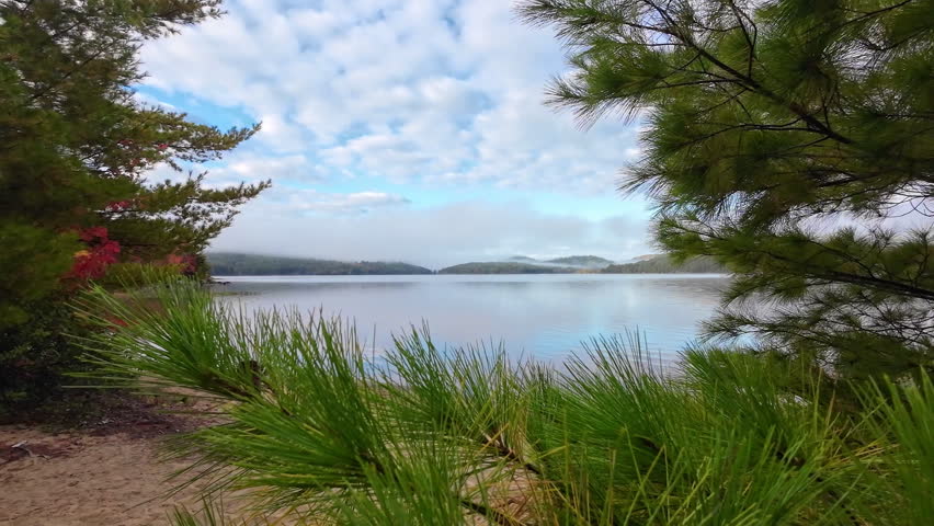 Slow motion reveal of a misty lake with sandy beach, forested hills and low clouds on a calm autumn morning in Algonquin Provincial Park.