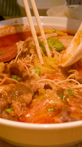 Close-up shot of chopsticks lifting beef and noodles from a spicy red broth bowl.