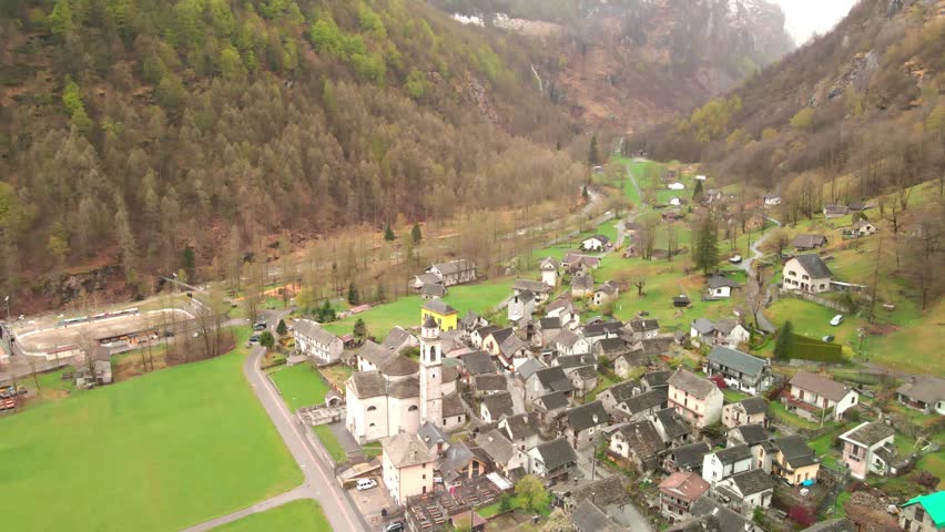 Aerial drone footage showcasing quaint rural village nestled in deep mountain valley, European landscape with traditional stone buildings, church steeple, winding roads and foggy peaks.