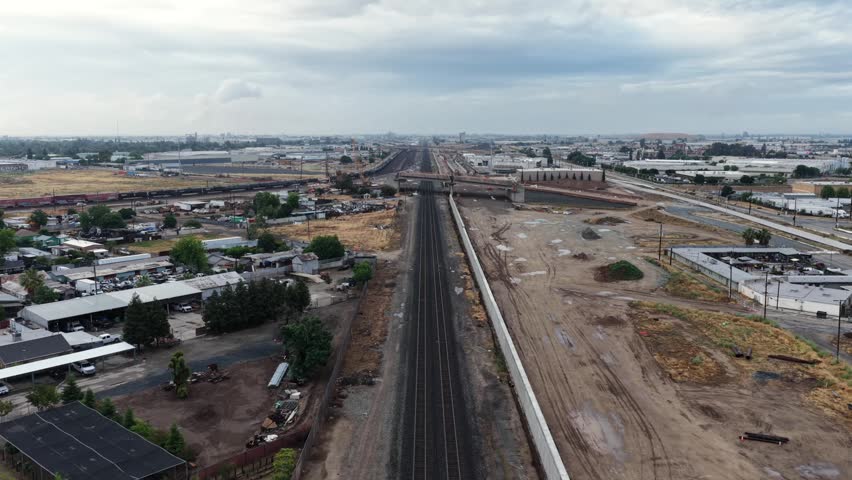 Empty rail corridor stretches between industrial warehouses and dirt lots, sprawling cityscape fading toward distant horizon beneath overcast sky. Fresno, California, USA. Aerial drone Birds Eye shot
