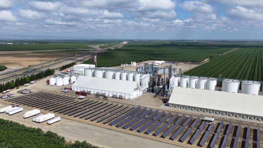 Almond processing facility in Arbuckle features white storage silos, solar panels, truck staging, and irrigated orchards stretching across Central Valley. California, USA. Aerial drone wide shot