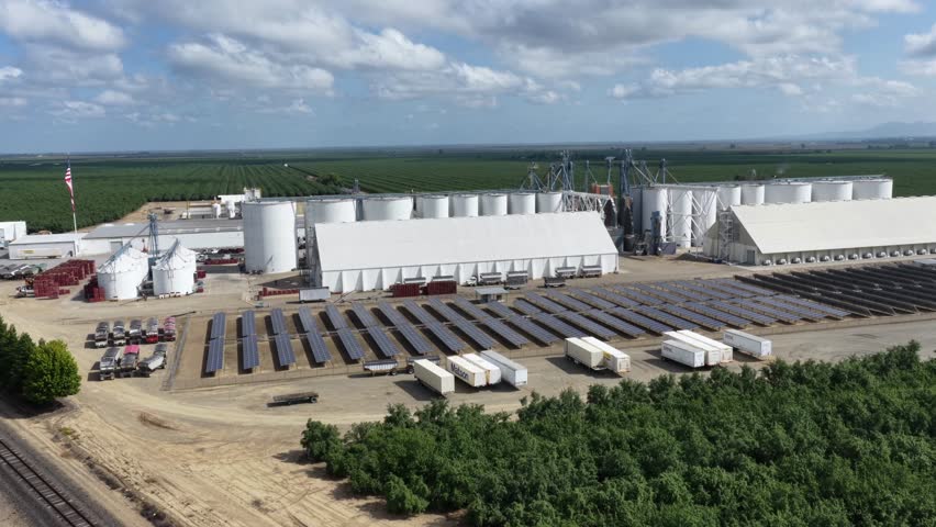 Almond processing facility with grain silos, solar panels and warehouse buildings surrounded by vast green orchard rows under dramatic clouds in Arbuckle, California, USA. Aerial drone rise wide