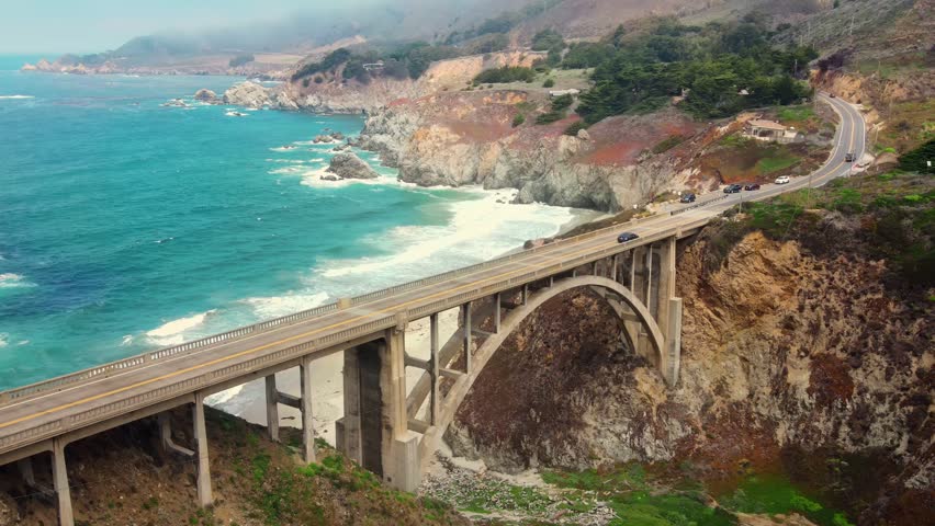 Cinematic aerial drone footage capturing Bixby Creek Bridge spanning rugged coastline in Big Sur California. Stunning coastal scenery featuring turquoise ocean waves Pacific Highway.
