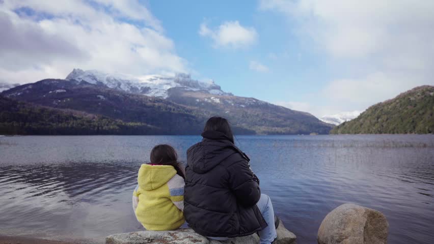 Mother and daughter drinking mate on the shore of Lake Falkner, Neuquen, Argentina, with snow-capped Andes mountains and Patagonian forest reflected in calm water, static lakeside shot.